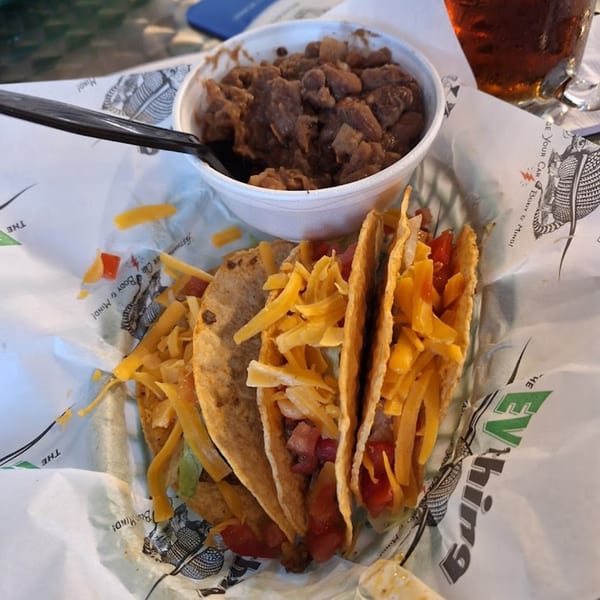 Three tacos with cheese and tomatoes, served with a side of beans and a drink.